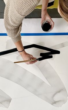 a woman is painting on the floor with white paint and black tape while another person holds a brush