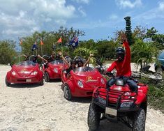 three people riding on four wheelers with flags flying in the air and two cars behind them