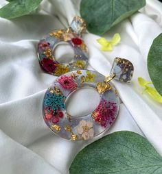 two colorful earrings sitting on top of a white cloth next to green leaves and flowers