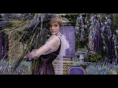 a woman in a purple dress is holding lavenders and looking at the camera with her hands behind her back