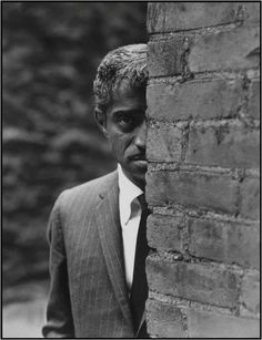 a black and white photo of a man in a suit leaning against a brick wall