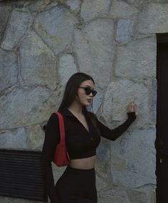 a woman in black outfit standing next to a stone wall with sunglasses on her head