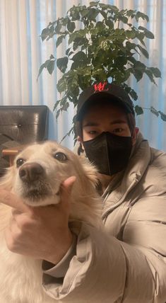 a man wearing a face mask holds his dog in front of him with a potted plant behind him