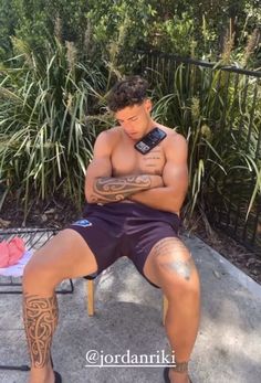 a shirtless man sitting on a chair looking at his cell phone while wearing tattoos