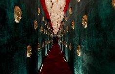 the hallway is lined with green walls and red carpeted flooring that has gold masks hanging from the ceiling