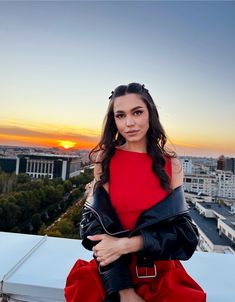 a woman in a red dress and black leather jacket posing for a photo on top of a building