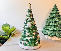 two ceramic christmas trees sitting on top of a white table next to a green plant