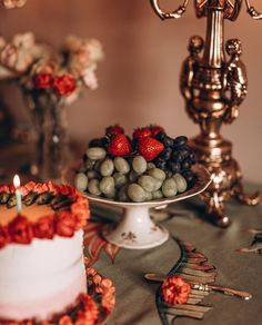 a table topped with a white cake covered in berries and grapes next to a gold candelabra