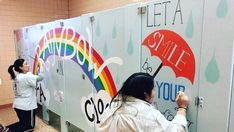 two women are painting the side of a bathroom stall with umbrellas and rainbow signs on it