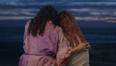 two women standing next to each other in front of the ocean at night with their arms around each other