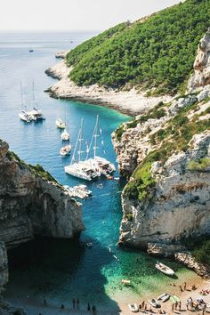 several boats are docked in the blue water near some cliffs and people on the beach