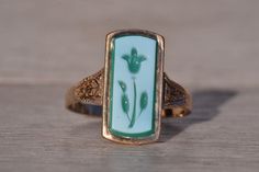 The Lynnfield: Antique Floral Carved Cameo Ring in Yellow Gold. This striking ring showcases an elongated tablet of chalcedony, adorned with a delicately hand-engraved flower motif in white and green hues, securely set within a flush bezel. On either side of the center, intricate hand-engraved patterns adorn the top of the shank, adding a touch of intricate detail to the design. Crafted with care in yellow gold, the ring exudes warmth and elegance. Currently sized at 9.25. Victorian Rings Engagement, Rich Wishlist, Green Ring Vintage, Edwardian Ring, Engraved Flower, Antique Floral, Cameo Ring, Green Hues, Cabochon Ring