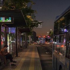 a man sitting on a bench next to a bus stop at night with city lights in the background