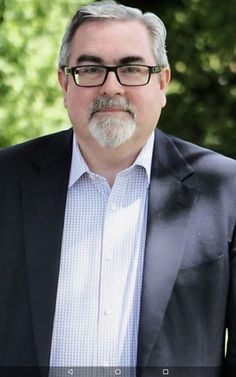 a man with glasses and a beard wearing a black suit jacket standing in front of trees