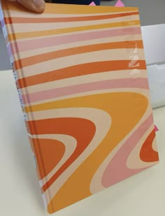 an orange and pink book being held up by a person's hand on a white table