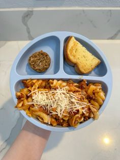 a person holding a blue plate with pasta and bread in it on top of a table