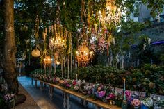 a long table covered in lots of flowers and hanging lights next to a lush green forest