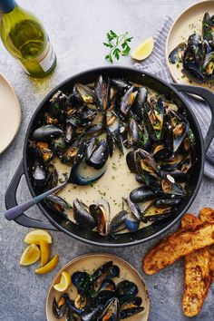 a pan filled with mussels and sauce next to two plates of bread on a table