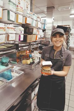 a woman in an apron is holding a sandwich and smiling at the camera while standing next to a counter