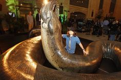 a woman standing next to a giant snake statue in a building with people looking on