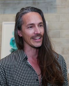 a man with long hair wearing a black and white checkered shirt smiling at the camera