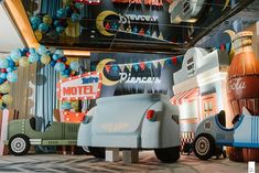 an assortment of toy cars and trucks are on display in a building with balloons hanging from the ceiling