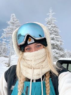 a woman with long hair wearing a blue ski helmet and scarf in front of snow covered trees