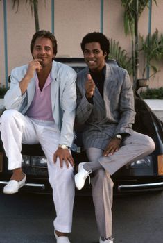 two men in suits sitting on the back of a black car and giving thumbs up