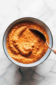 a hand holding a spoon over a bowl of carrot hummus on a marble surface