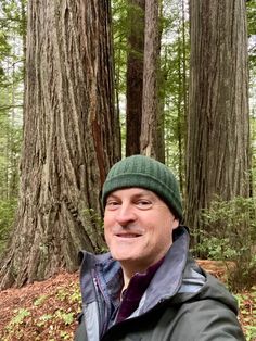 a man wearing a green hat standing in front of some tall tree trunks and trees