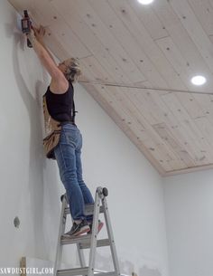 a woman on a ladder painting the ceiling in a room with white walls and wood planks