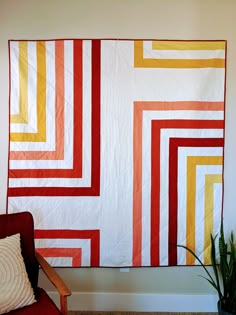 a red chair sitting in front of a white wall with an orange and yellow quilt on it