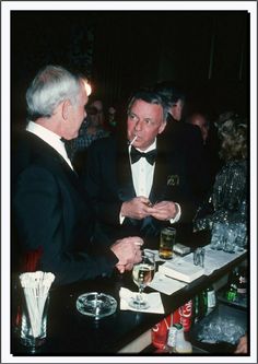 two men in tuxedos sitting at a bar talking to each other and drinking