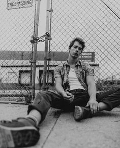 a young man sitting on the ground next to a chain link fence with his legs crossed