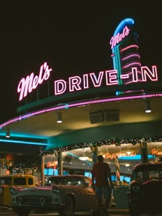 cars parked in front of a drive - in with neon lights on the building and people walking by