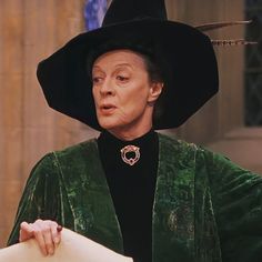 an older woman wearing a green velvet outfit and black hat holding a large piece of paper