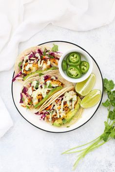 three tacos on a plate with limes and jalapenos next to them