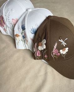 three baseball caps with embroidered flowers on them sitting on a white bedding sheet, one has a dragonfly and the other has an insect