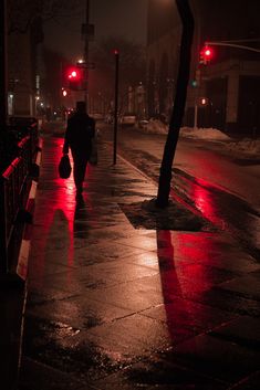 a person walking down the street in the rain at night with red traffic lights on