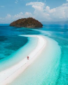 two people are walking on the beach in front of an island and clear blue water