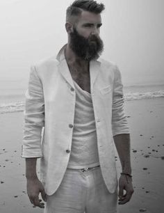 a man with a beard and white suit standing on the beach in front of the ocean