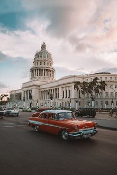 an old red car driving down the street in front of a building with a dome