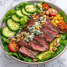 a steak salad with tomatoes, cucumbers, and blue cheese dressing in a white bowl on a marble surface