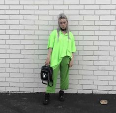 a woman standing in front of a white brick wall holding a black bag and wearing green pants