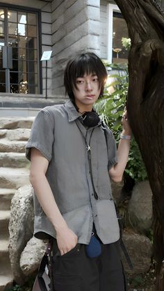 a young man standing in front of a tree with his hand on his hip, wearing a gray shirt and black pants