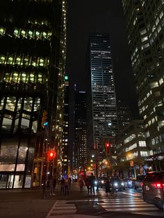 people are crossing the street at night in front of tall buildings and skyscrapers with traffic lights