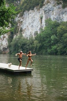 two people jumping into the water from a dock in front of a mountain side cliff