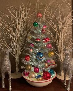 a small christmas tree in a bowl with ornaments around it and two deer figurines next to it