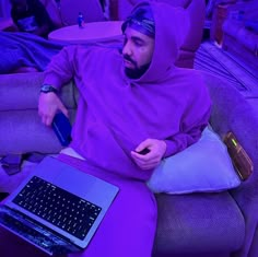 a man in a purple hoodie sitting on a couch with a laptop and cell phone