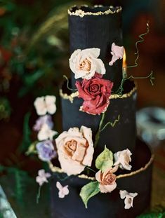 a three tiered black cake with flowers on it's side and a lit candle in the middle